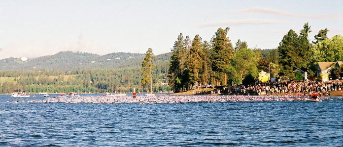 [There is a wide swatch of swimmers from the shore to quite a ways out in the water. Lots of color and flying water in the lake with the tall trees on shore catching the morning sun.]